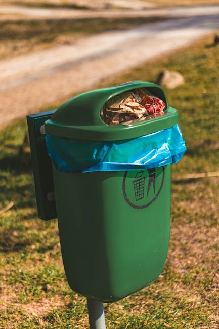A green outdoor rubbish bin with a rounded, dome-shaped lid is positioned on a grassy area, attached to a metal pole. The bin's lid is partially open, revealing a clear plastic bag filled with mixed waste, including crumpled paper, plastic packaging, and foil wrappers. A blue plastic bag lines the interior of the bin, secured around the top edge. The bin displays a black graphic icon of a trash can within a circle, indicating waste disposal. The background shows a blurred outdoor scene with a dirt pathway and patches of grass, suggesting a park or communal outdoor space. The environment appears well-lit, with natural sunlight casting minimal shadows. The scene exemplifies a typical location for waste collection, potentially associated with private or independent rubbish disposal services, as managed by Waste Collection Belsize Park, in accordance with the local rubbish removal needs.
