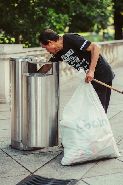 A green outdoor rubbish bin with a rounded, dome-shaped lid is positioned on a grassy area, attached to a metal pole. The bin's lid is partially open, revealing a clear plastic bag filled with mixed waste, including crumpled paper, plastic packaging, and foil wrappers. A blue plastic bag lines the interior of the bin, secured around the top edge. The bin displays a black graphic icon of a trash can within a circle, indicating waste disposal. The background shows a blurred outdoor scene with a dirt pathway and patches of grass, suggesting a park or communal outdoor space. The environment appears well-lit, with natural sunlight casting minimal shadows. The scene exemplifies a typical location for waste collection, potentially associated with private or independent rubbish disposal services, as managed by Waste Collection Belsize Park, in accordance with the local rubbish removal needs.