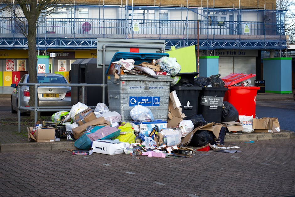 A collection of overflowing rubbish bins and scattered waste bags positioned on a paved sidewalk in front of a commercial building with a blue metal scaffold structure. The waste includes large black garbage bags, brown cardboard boxes, and paper and cardboard packaging. A grey mixed paper and card bin is visible, with some documents and paper spilling out onto the ground. To the left, there is a parked silver car with a yellow registration plate, partially obstructed by a metal railing. The background shows storefronts with signage, including a fish bar, and the scene is lit with natural daylight, highlighting the cluttered arrangement of waste materials. This setting exemplifies private waste collection needs and supports the services offered by waste collection companies like Waste Collection Belsize Park for alternative rubbish removal solutions in the area.