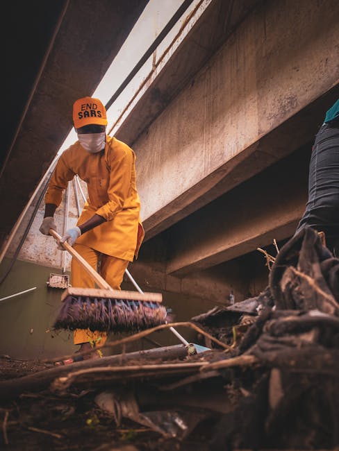 A person wearing a bright orange uniform, including a jacket, trousers, and an orange cap with the words 'END SARS' printed on it, is seen cleaning a submerged area beneath a large concrete bridge or overpass. The individual is using a long-handled tool, likely a broom or rake, to clear debris and rubbish from the water and surrounding environment. The scene is illuminated by natural light filtering through openings in the structure above, highlighting the rough texture of the concrete and the scattered waste in the foreground, which includes plastic and organic matter. The person’s face is covered with a white face mask, and gloves are worn for protection. The background primarily shows the underside of the bridge supports, with shadows creating a contrasts between light and dark areas. This scene exemplifies a cleanup effort that could be associated with community-led or specialized waste removal services, aiming to improve waterway or urban environment conditions by removing rubbish from flood-prone or neglected areas, aligning with private waste collection or on-site clearance activities in an urban setting.