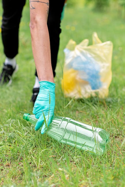 A person wearing a light blue disposable glove is seen picking up a clear plastic bottle from a grassy outdoor area. In the background, there is a yellow and blue plastic bag resting on the ground, partially filled with other waste items. The scene appears to be part of a litter collection activity, with the person focused on removing waste from the environment to support effective rubbish management. The grass is green and slightly uneven, indicating an outdoor natural space, and the lighting suggests daytime with natural light. This image illustrates environmental cleanup efforts and aligns with waste removal services, such as those provided by Waste Collection Belsize Park, emphasizing the importance of proper waste disposal and community engagement in maintaining clean surroundings.